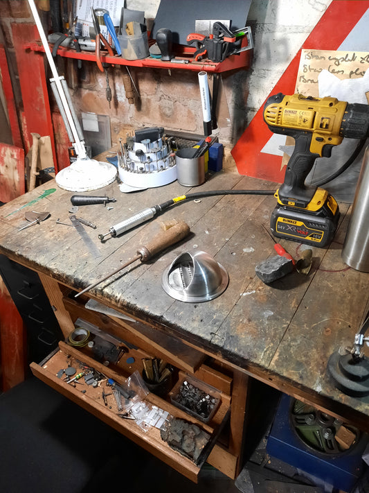 Looking down from top left at a Messy jewellers workbench. Tools, polishing compoumd, lamp, strewn over the bench. A round, domed, stainless steel ventilation duct in the middle used for extraction.